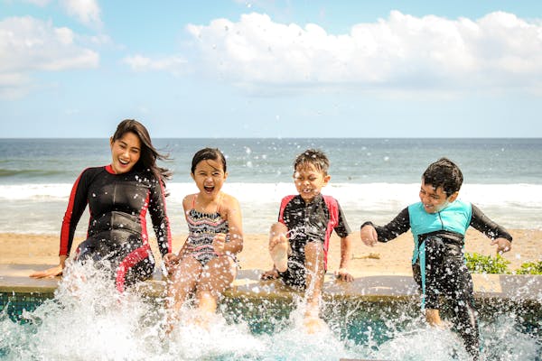A joyful group of children and an adult splashing water while sitting by a pool with a beach view.