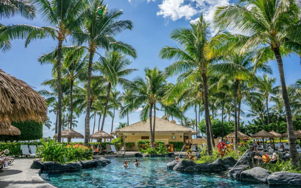 A tropical resort pool surrounded by palm trees, vibrant flowers, and thatched-roof cabanas under a sunny sky.