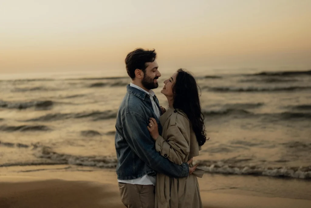 A couple embracing on the beach at sunset, gazing into each other's eyes with the ocean waves in the background.