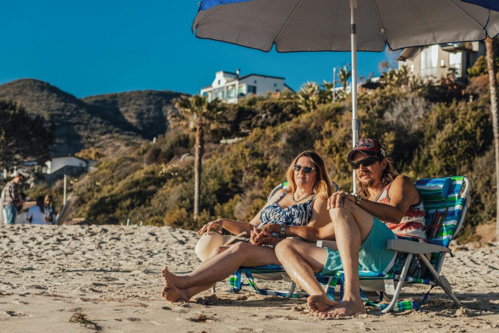  A couple sitting under an umbrella on the beach, relaxing in beach chairs with a scenic hillside and houses in the background.
