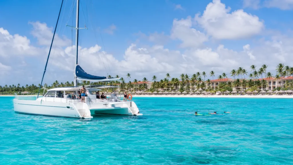 White catamaran tour boat on turquoise Punta Cana water with snorkelers, palm trees, and resort coastline in the background