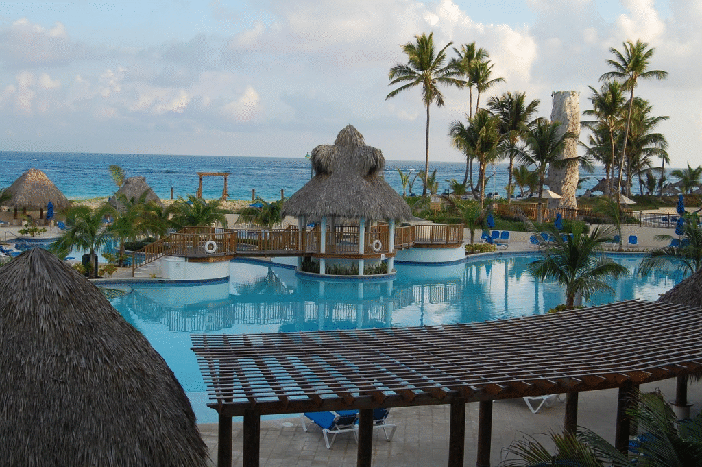 A serene resort pool area with thatched-roof cabanas, palm trees, and a view of the ocean under a partly cloudy sky.