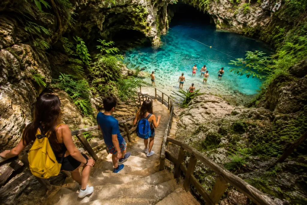 Family walking down stone steps to a turquoise cenote at Scape Park in Punta Cana, with people swimming below.