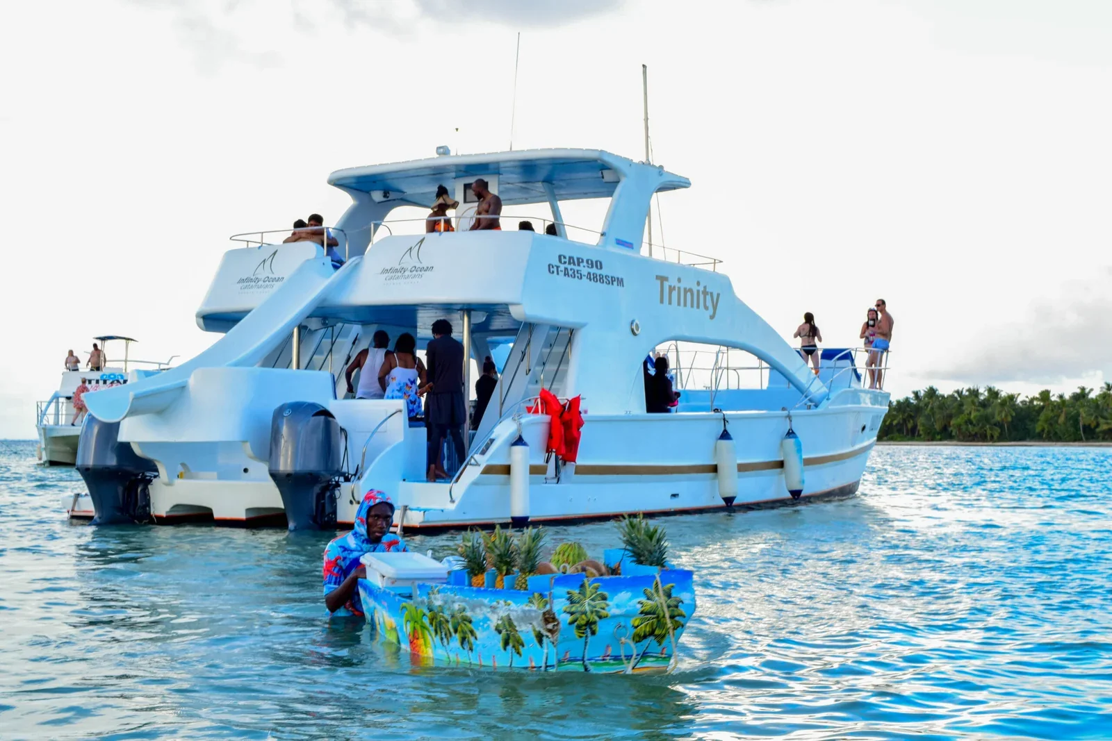 A boat named "Trinity" in clear waters, with people on deck and a pineapple vendor approaching during a Catamaran Tour Punta Cana.