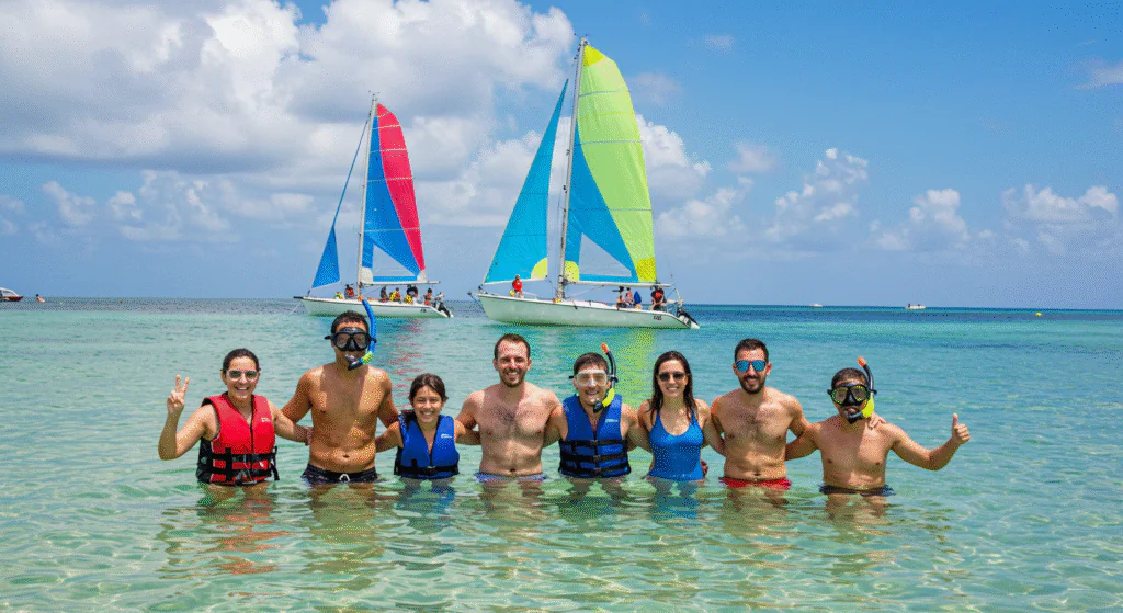 A group of friends and family in life vests smiles and poses in shallow water, with colorful sailboats in the background.